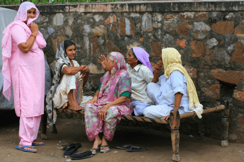 four women sitting on charpai and talking while a fifth woman stands and listens